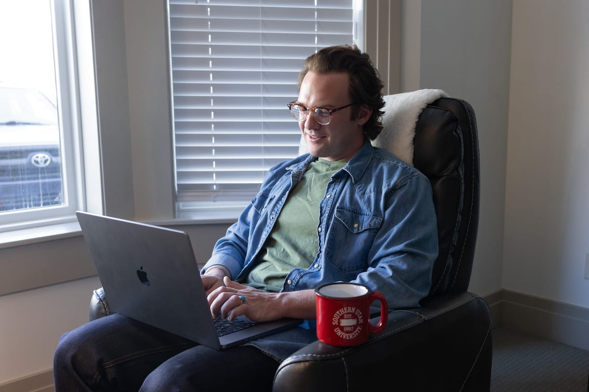 An adult online student sitting on a chair with a laptop on his lap, and a red cup with SUU sign next to him.