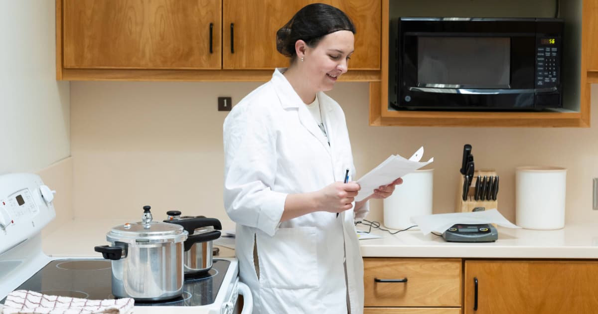 A student is taking a nutrition science class in the kitchen with some papers and a pencil in her hand, and some cookware in the background.