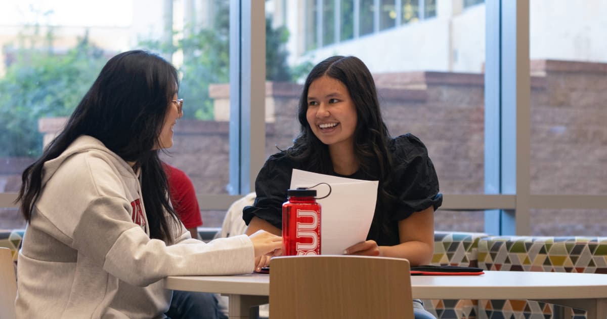 Two students sitting at a table chatting together. One student is holding a piece of paper, likely an essay or other piece of schoolwork. There is a red S.U.U. water bottle on the table. 