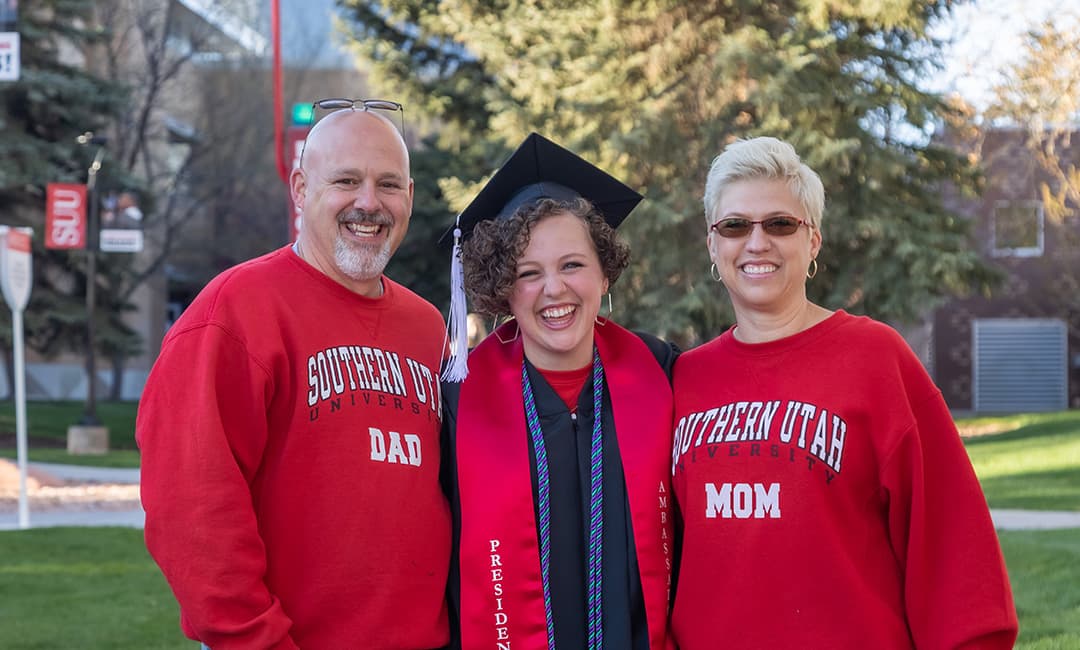 A smiling SUU student in graduation regalia standing between her parents