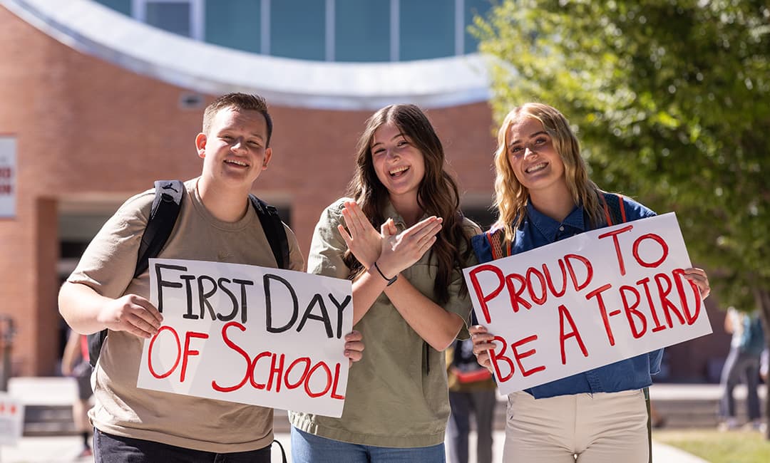 New SUU students holding signs in front of the library that read 'First Day of School' and 'Proud To Be A T-Bird'