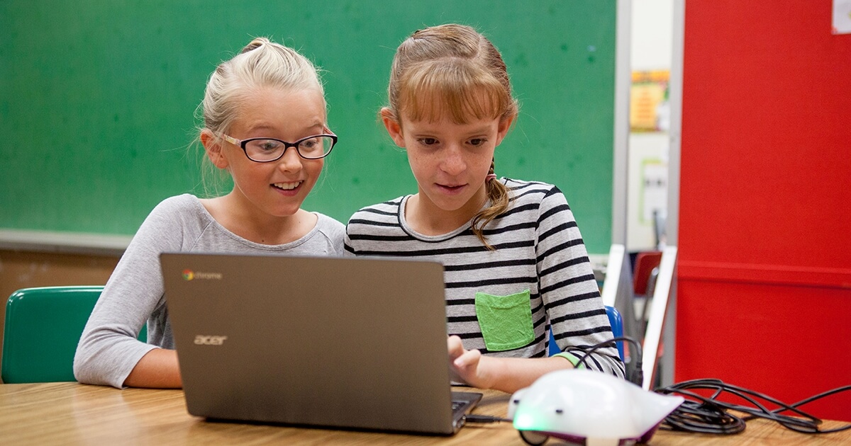 Two young girls working on a laptop that is connected to a small robotic device