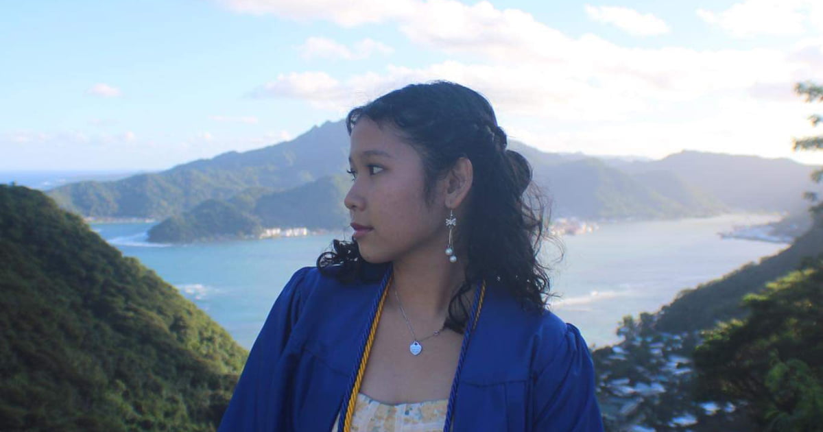 Photograph of Visca standing in her high school graduation gown looking away from the camera, an island view in the background.