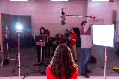 A behind-the-scenes photo of an interview being filmed. The back of a woman's head is seen in the foreground with students operating filmmaking equipment in the background.