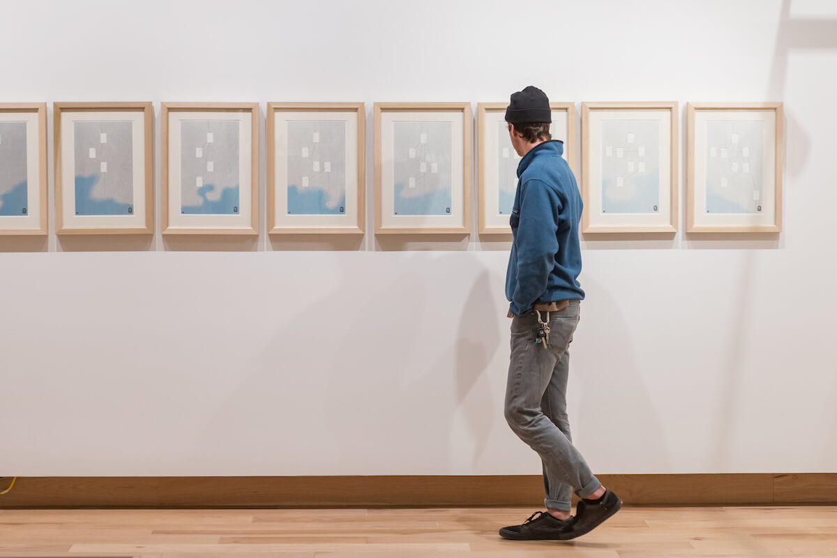 A man in a blue sweater looking at a series of artworks displayed next to each other in a line.